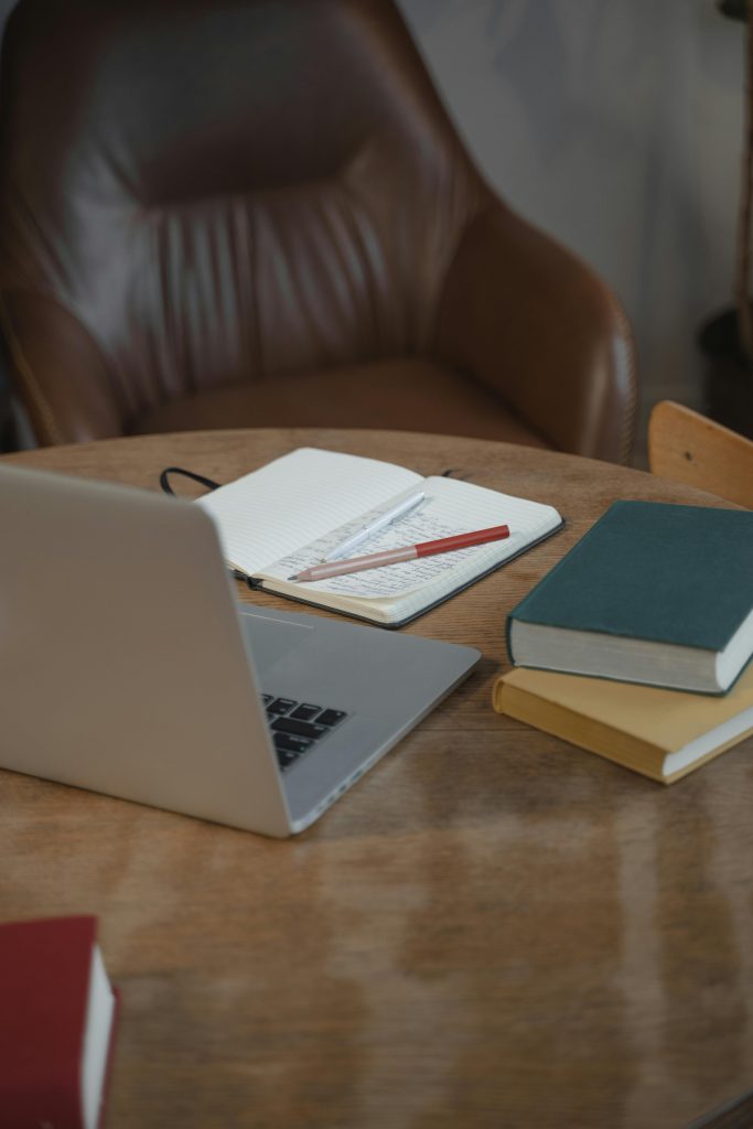 Ordenador portátil sobre una mesa, también se ven unos libros cerrados y una libreta abierta a la mitad con unas notas y unos bolígrafos